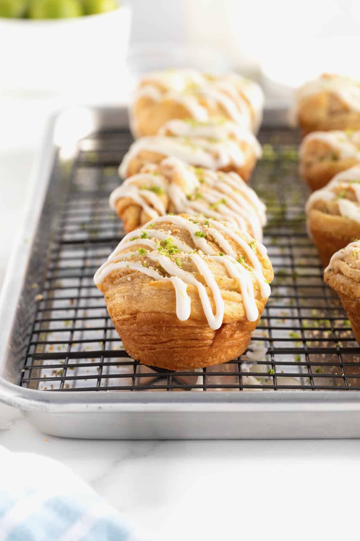 An aluminum baking pan with a wire cooling rack on top holding key lime cruffins garnished with lime zest and graham cracker crumbs.