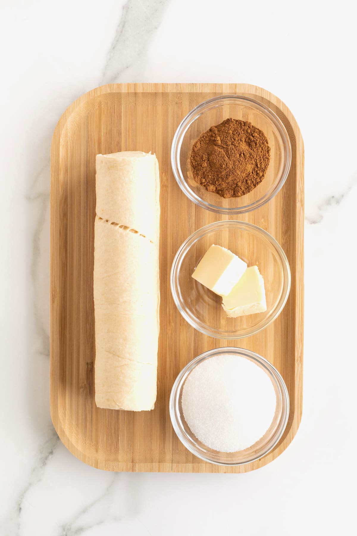 A roll of canned crescent dough and butter, cinnamon, and granulated sugar in small glass dishes on a rimmed wooden cutting board.