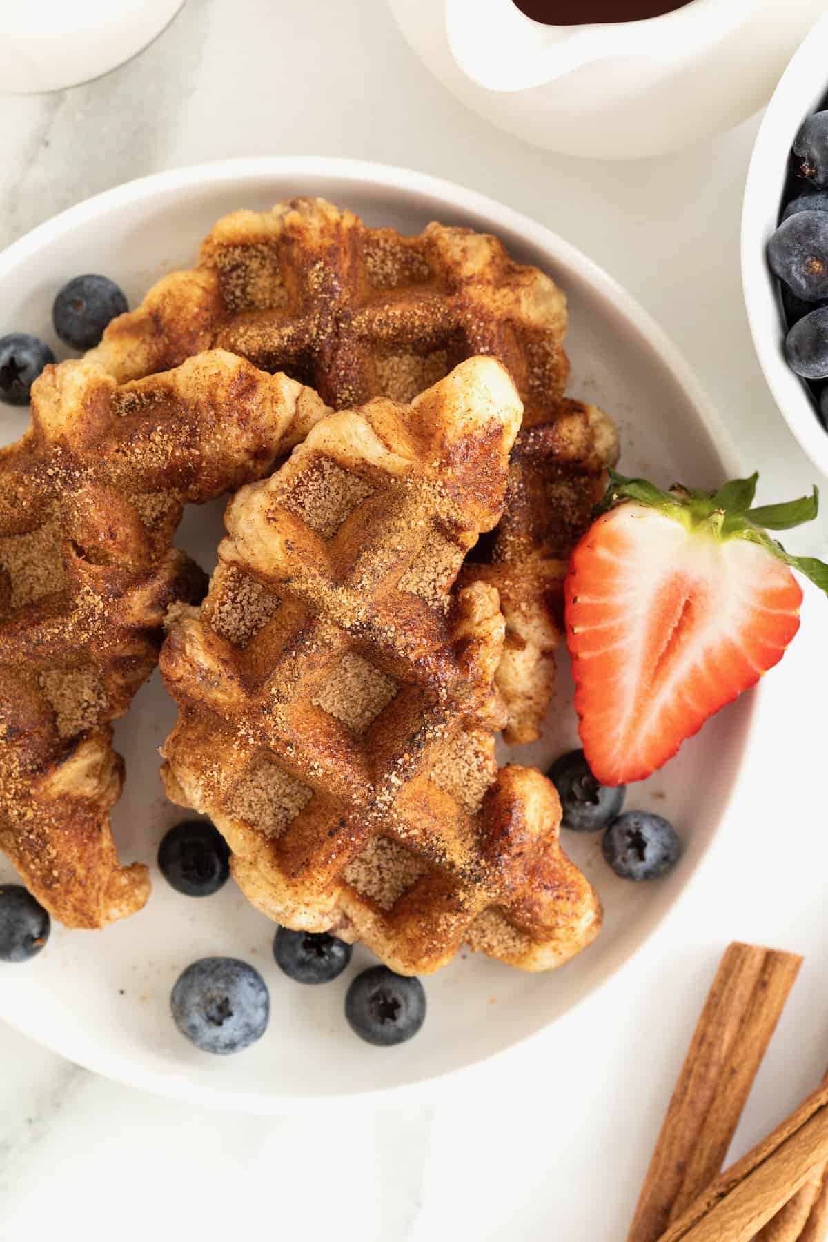 Cinnamon sugar croffles on a white rimmed plate surrounded by several blueberries and a strawberry half.