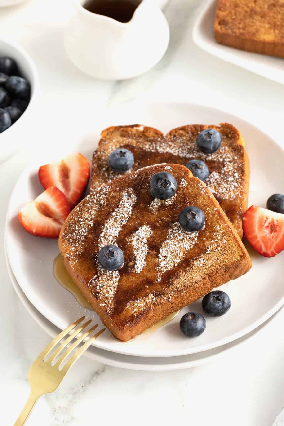 A white plate of three pieces of baked French toast topped with maple syrup and berries.
