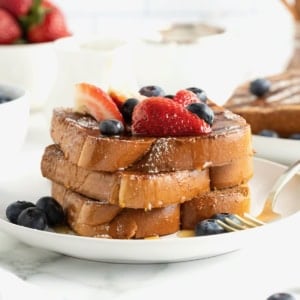 Three slices of baked French toast stacked on a white plate. The French toast is topped with blueberries, strawberry slices and dusted with powdered sugar.