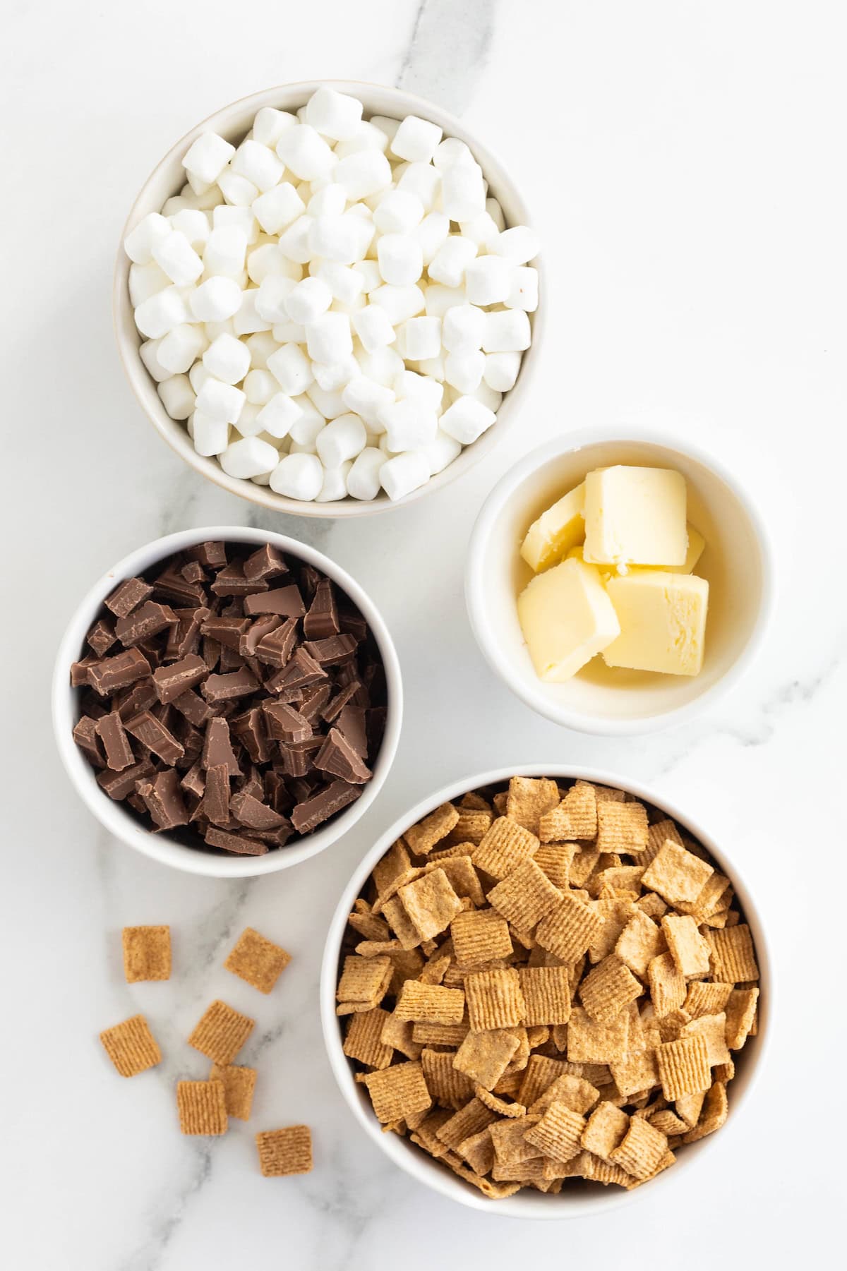 Ingredients to make No-Bake S'mores Treats in small white glass dishes on a white marble counter.